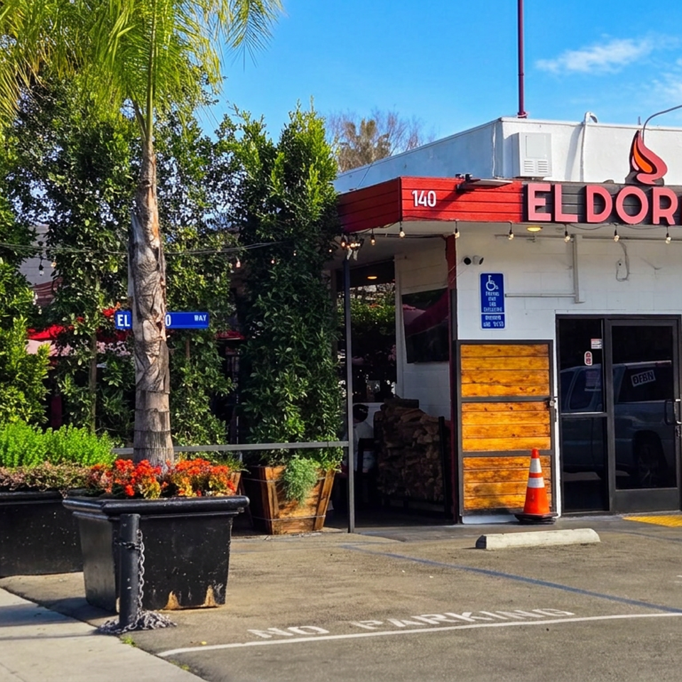 Sunny street view of Eldorado Pollo Asado restaurant entrance with tropical plants, outdoor seating, and accessible parking.